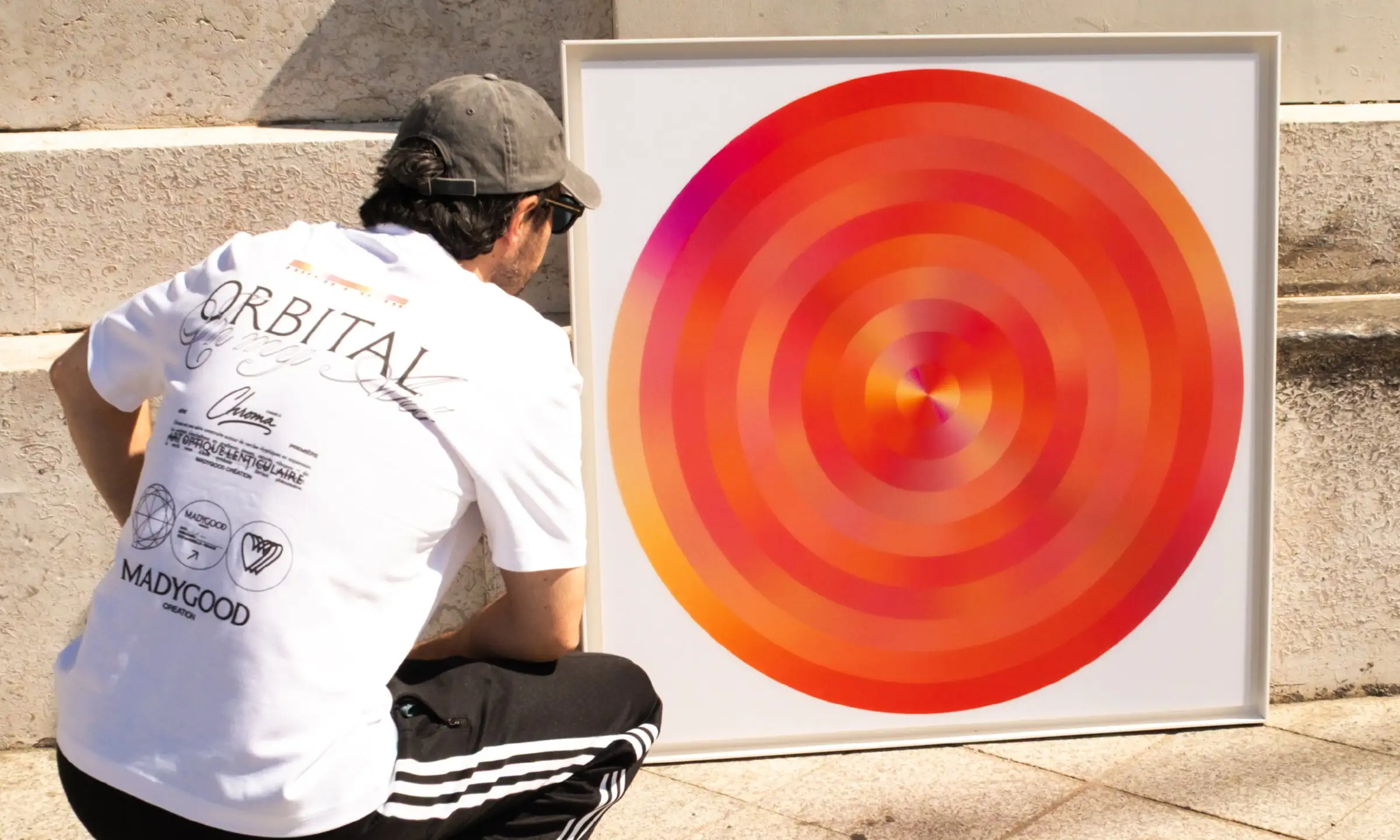 Anthony Celerier crouching, seen from behind in an ORBITAL t-shirt, facing a circular orange artwork in a white frame against a stone wall.