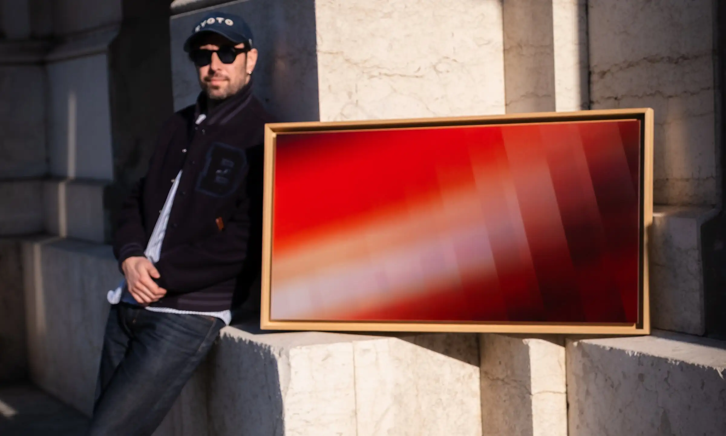 Anthony Celerier leaning against a marble wall, beside a red artwork in a wooden frame.