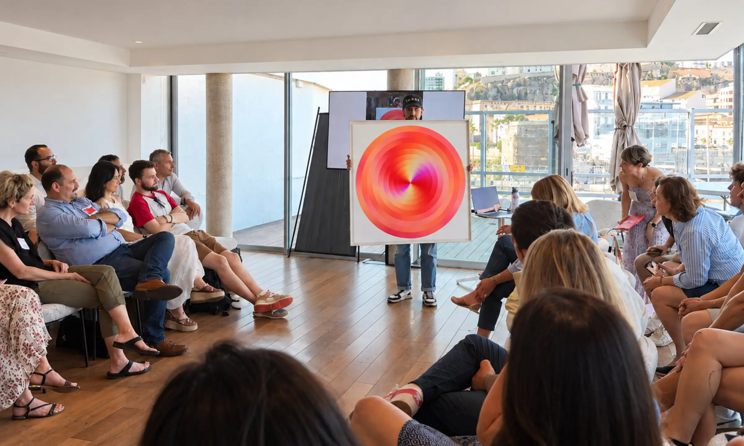 Anthony Celerier presents a circular orange artwork to an audience, in a room opening onto the sea.