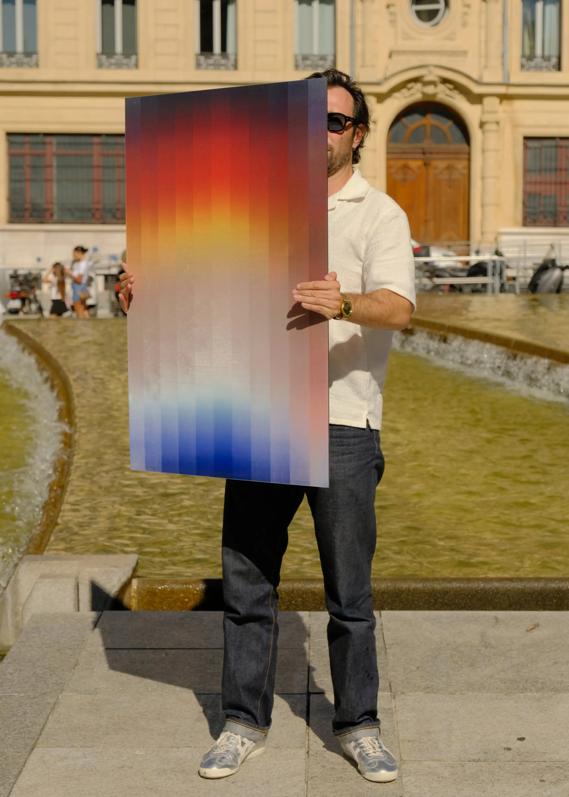 Anthony Celerier, wearing a cap and sunglasses, holds the white frame of an artwork in which an orange disc can be glimpsed, in front of a stone wall.