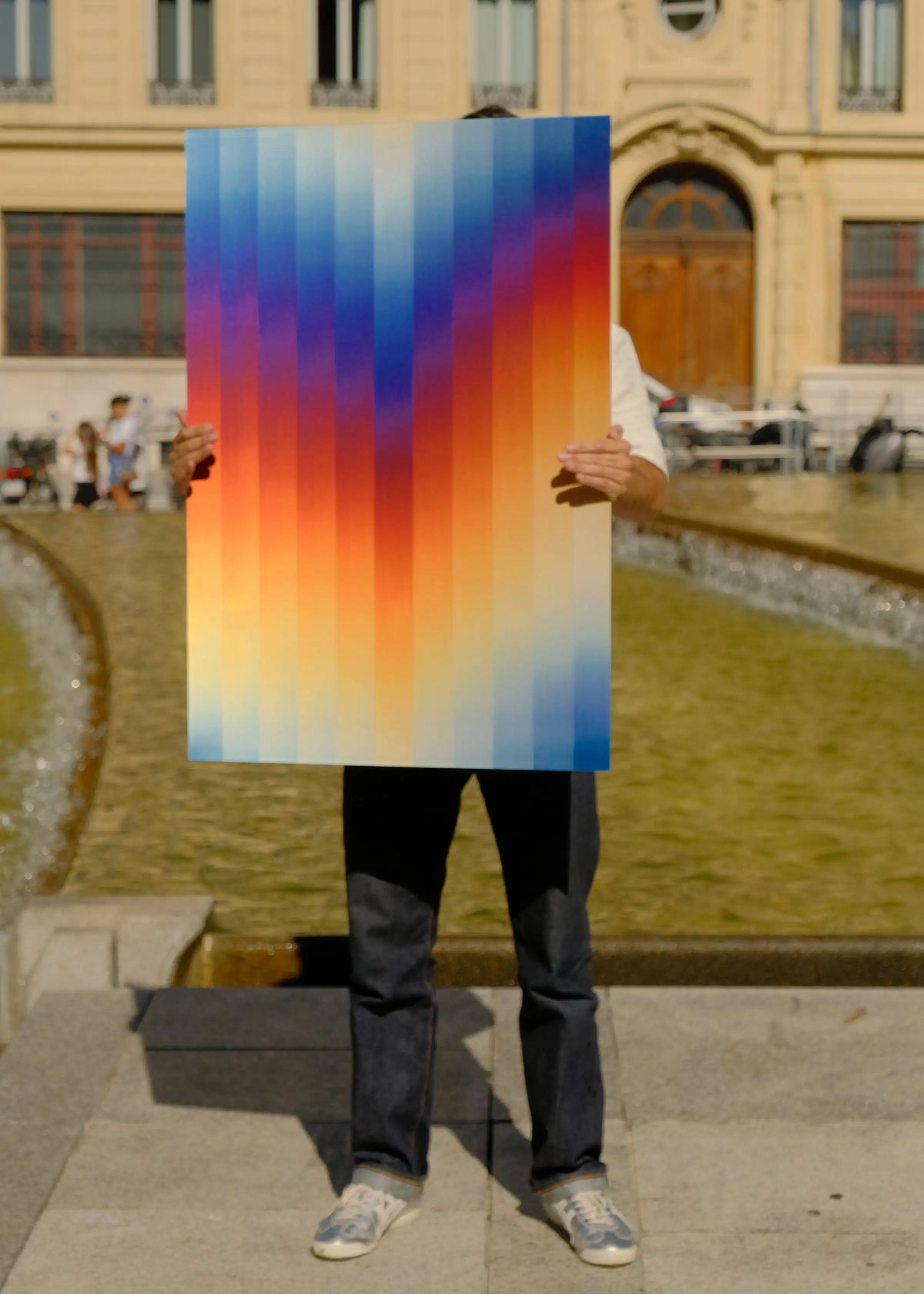 Anthony Celerier présente l’œuvre Sunrise, des lames de couleur dans un cadre, devant une fontaine.